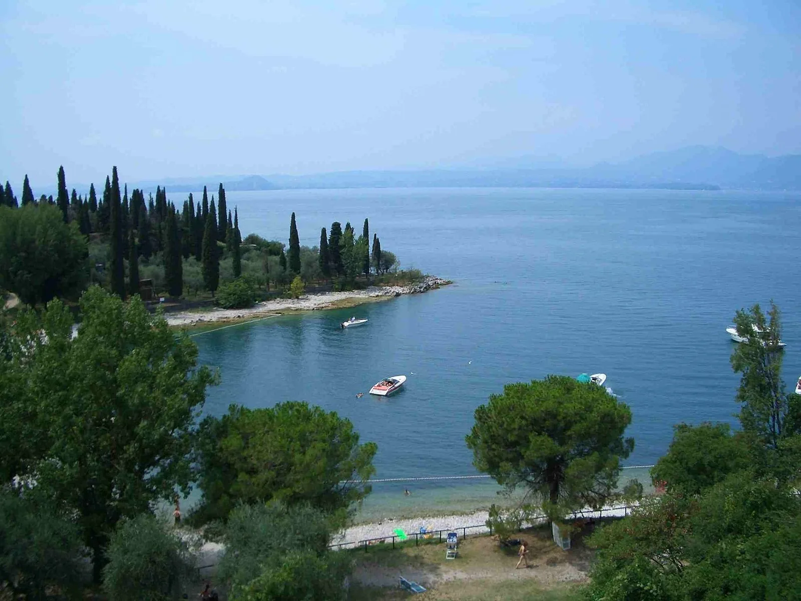 Baia con spiaggia di ciottoli, cipressi e barche sull'acqua blu del Lago di Garda in estate